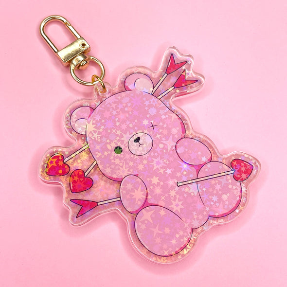 Acrylic keychain with star holographic finish and gold key ring. Artwork shows a super cute pink teddy bear missing an eye button with heart arrows through it.