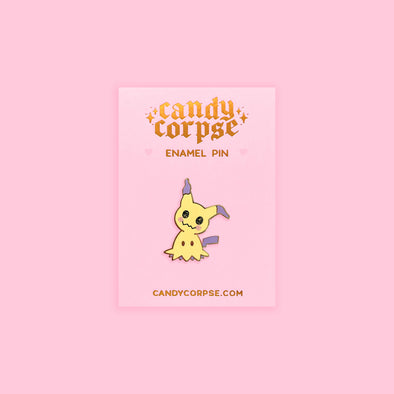 Enamel pin of a cute yellow monster playing dress up. Sitting on a pink backing card with gold foil Candy Corpse logo.