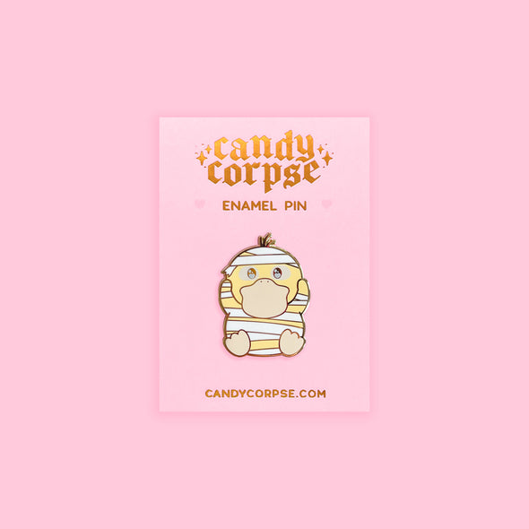 Enamel pin of cute duck monster dressed as a mummy for Halloween. Sitting on pink backing card with gold foil embossed Candy Corpse logo.