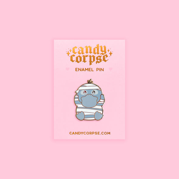 Enamel pin of cute duck monster dressed as a mummy for Halloween. Sitting on pink backing card with gold foil embossed Candy Corpse logo.