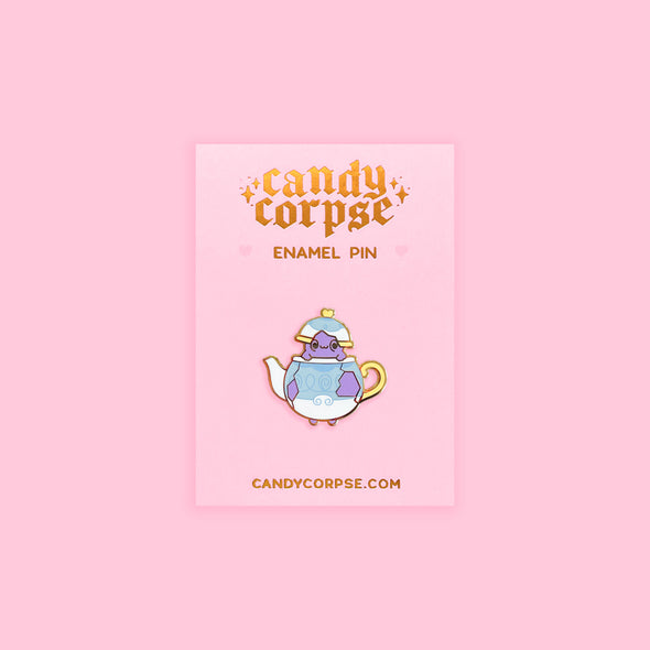 Enamel pin of a ghost in a teapot. Sitting on a pink backing card with gold foil Candy Corpse logo.